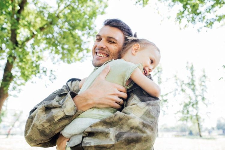 A service member in uniform hugging a young child outdoors