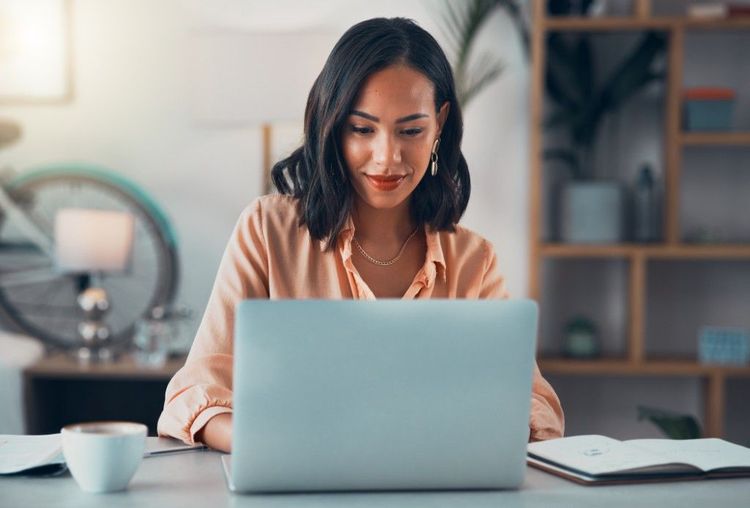 A woman working on a laptop at a desk with a notebook and coffee mug